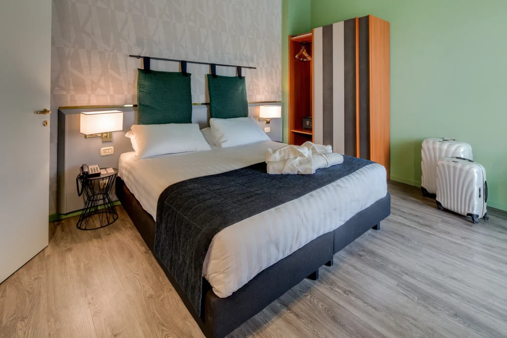 In the picture a room of hotel. On the left there is a double bed with a blue runner and two folded bathrobes on it. On the left there is a small black table with a phone on it and night lights on both sides of the bed. On the right there is a wooden closet with a safe inside and two suitcases next to it. The floor is parquet.