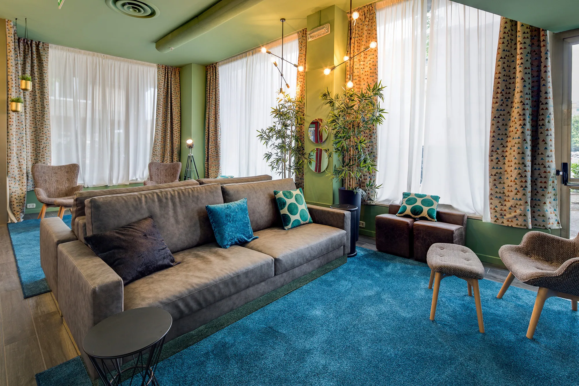 In the picture the hall of the hotel. In the center of the picture there are two sofas positioned against each other's backs. In front of them there are light brown armchairs and a stool. On the floor under the two different living spaces there is a blue rug. The walls are covered by windows and curtains.