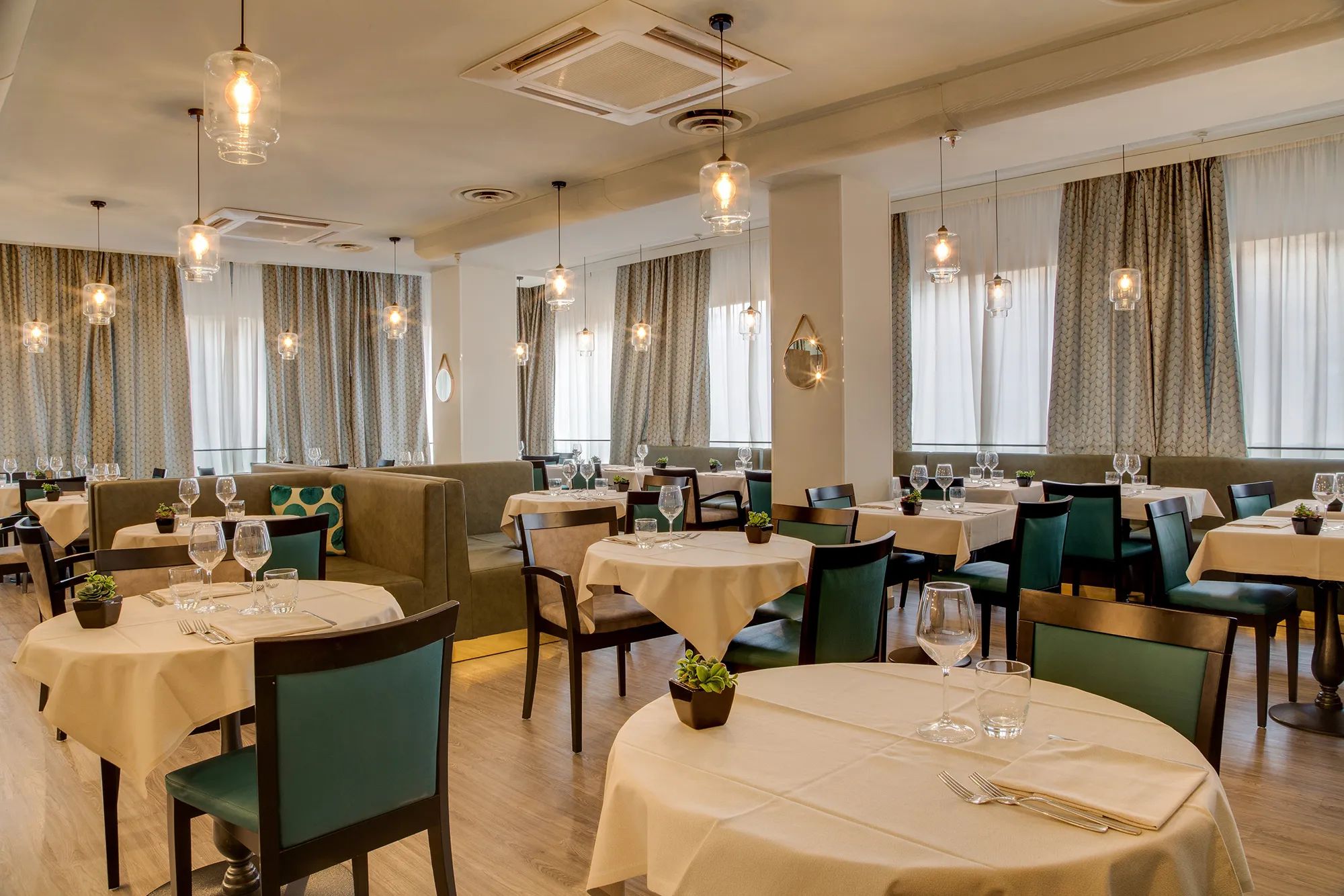 In the picture the restaurant of the hotel. There are many small tables with white tablecloth and beige and green leather and wood chairs. On the tables there are glasses, cutlery and napkins.