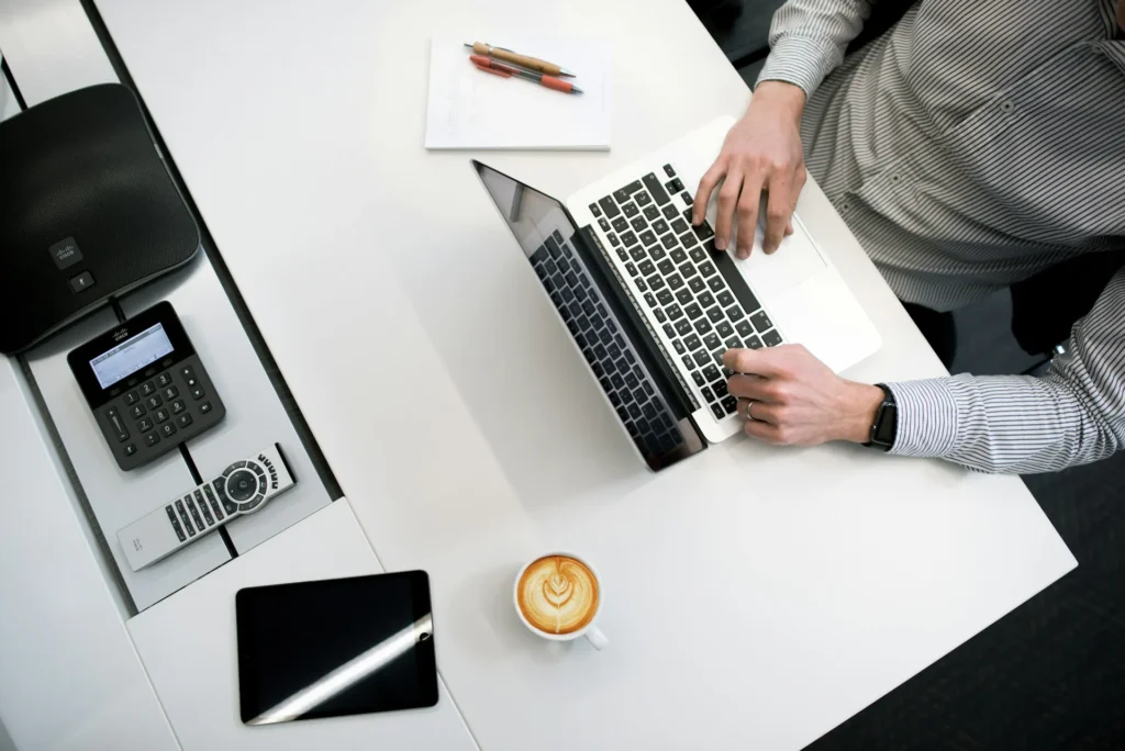 In the picture a man wearing a white and grey striped shirt is sitting on a chair and works on a laptop. Next to him there is a notebook and two pens. On the other side there is a cappuccino. On the left side of the table there is a tablet, a remote, a keypad and a sound box
