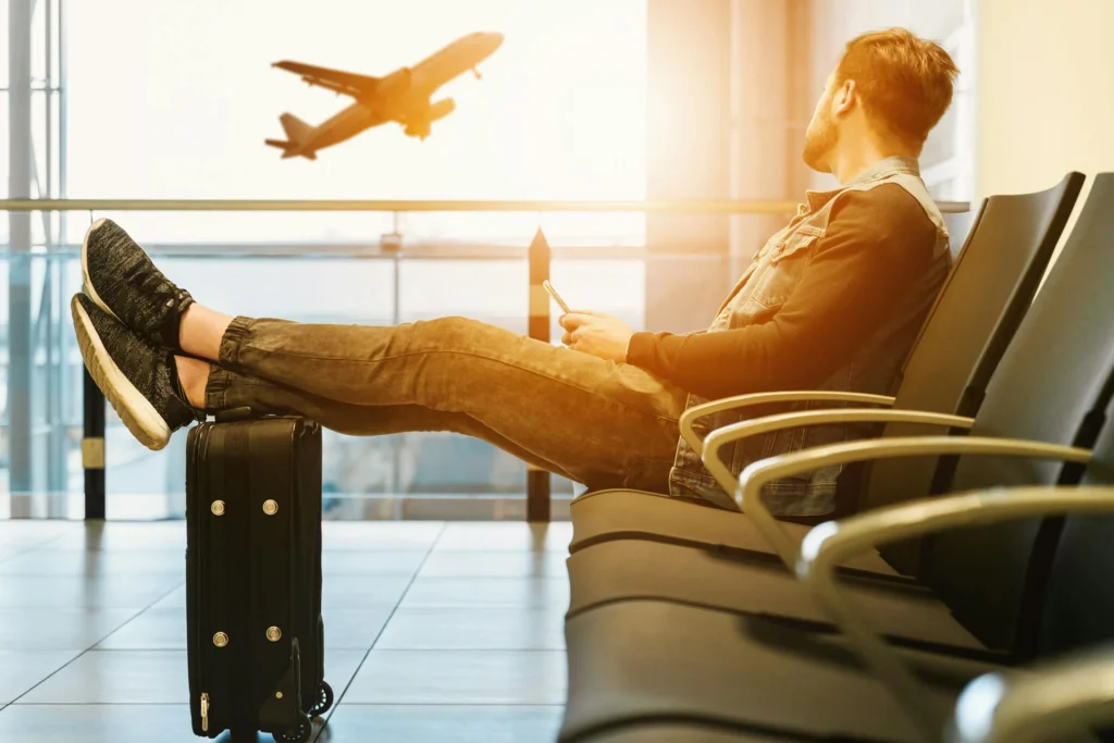 In the picture there is a men sitting on a plastic bench of the airport. His legs are stretched out on the suitcase. He is watching a plane landing out of the window.